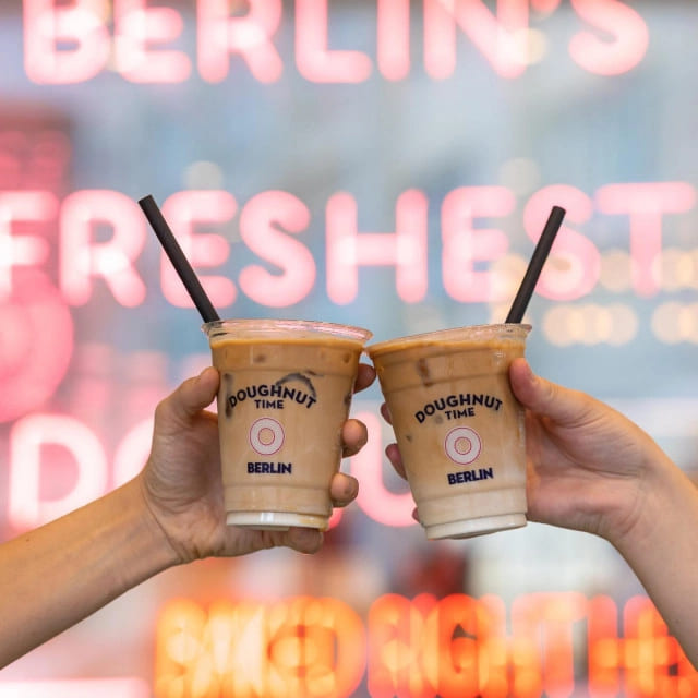 Two hands holding iced coffee drinks in branded plastic cups with Doughnut Time logo, set against a vibrant backdrop. Perfect example of branded plastic cups used for café promotions.